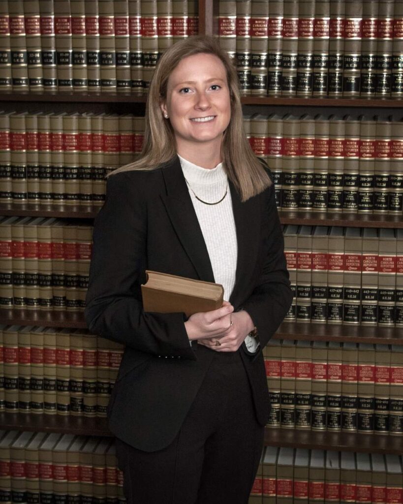Aerin in professional attire stands in front of a bookshelf filled with legal books, holding a book and smiling at the camera.