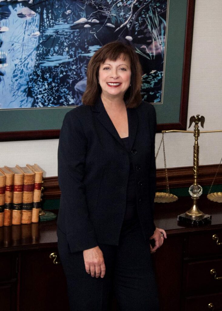 Deborah in a dark suit stands in front of a framed picture, next to a desk with books and a brass lamp.