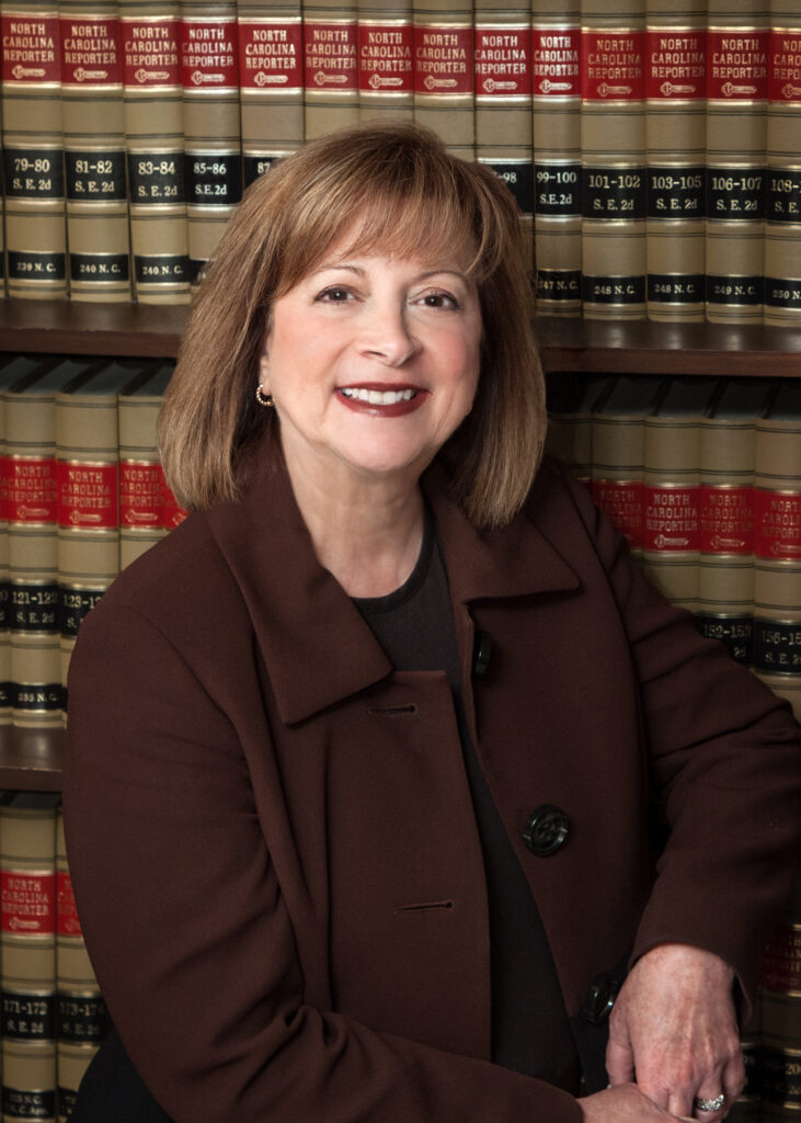 Deborah in a suit stands stands smiling in front of shelves filled with books.