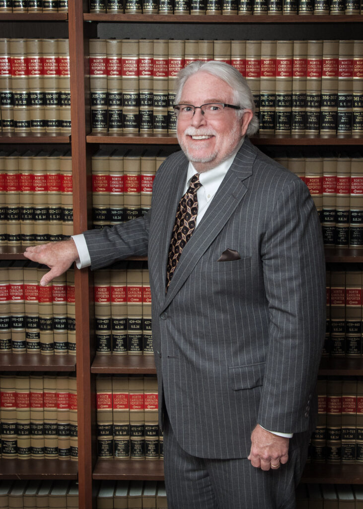 Paul in a gray suit and white shirt, standing confidently with one hand in his pocket in front of shelves filled with law books.