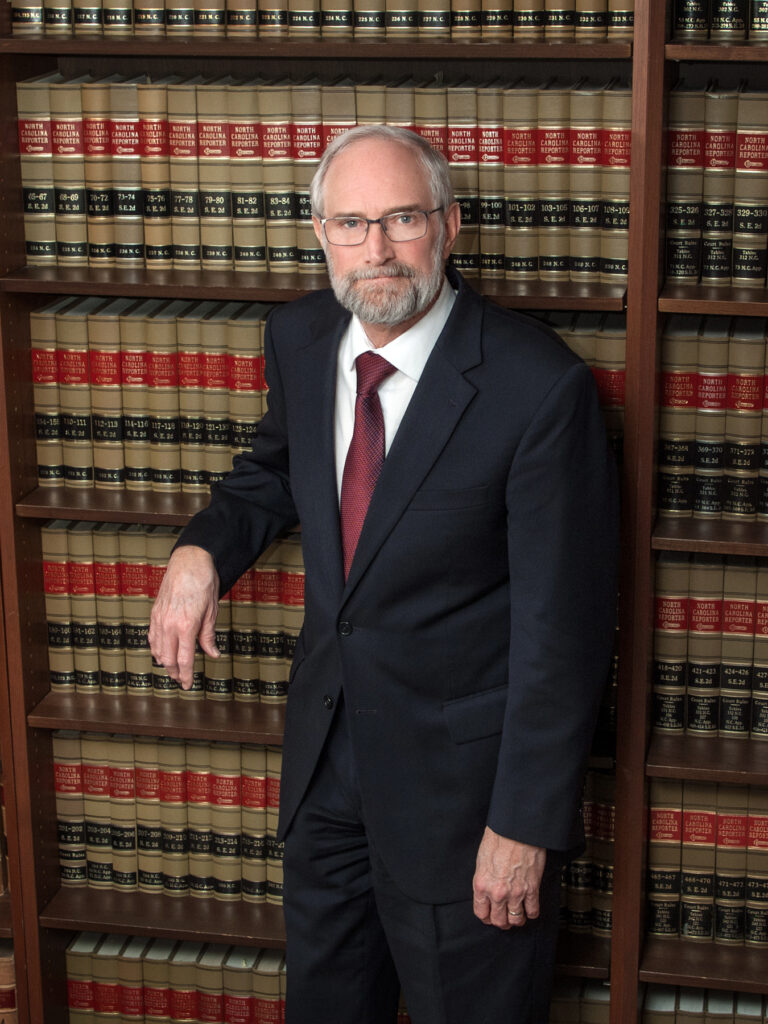 Ken in a dark suit and patterned tie stands in front of a bookshelf filled with legal books, one hand in his pocket.
