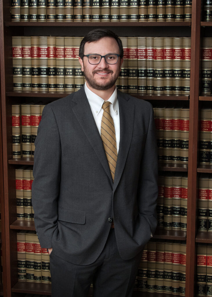 Andrew in a gray suit, white shirt, and yellow tie stands in front of bookshelves filled with law books.