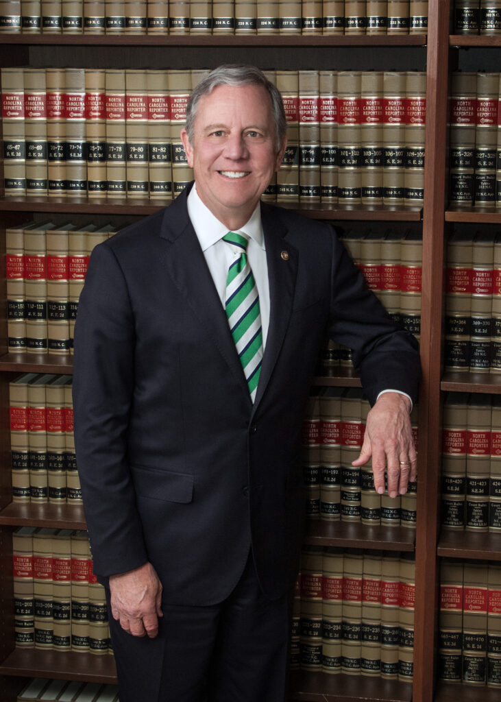 Rick in a dark suit and green striped tie stands smiling in front of shelves filled with books.