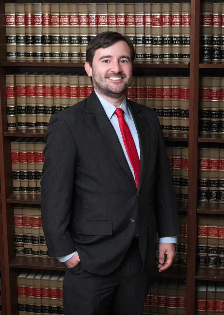 Adam with a beard, wearing a dark suit and red tie, stands in front of shelves filled with numerous books.
